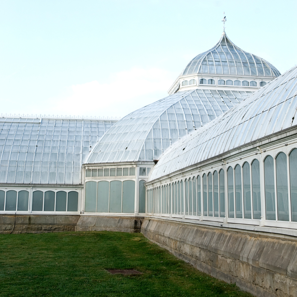 A close-up view of the intricate glass domes of the Phipps Conservatory and Botanical Gardens in Pittsburgh, showcasing the building’s historic architecture and large, curved windows against a backdrop of clear sky and well-manicured grass.