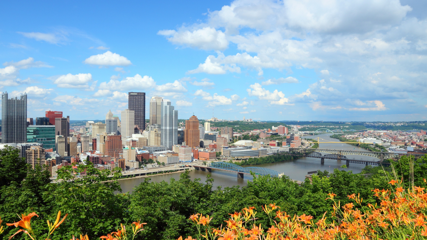 A bright and colorful view of downtown Pittsburgh on a sunny day, with the city's skyline framed by lush greenery and vibrant orange lilies in the foreground. The rivers and bridges that define the city's landscape stretch out under a blue sky dotted with clouds.