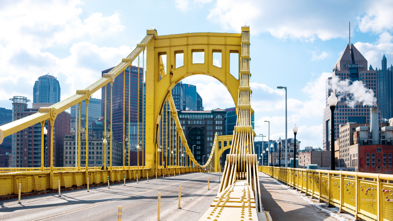 A close-up view of Pittsburgh's iconic yellow Roberto Clemente Bridge, looking toward the city's skyscrapers. The bright bridge structure contrasts with the cool blue sky and the buildings in the background.