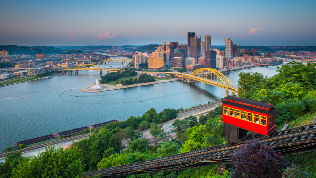 A scenic view of downtown Pittsburgh, featuring the confluence of rivers, iconic yellow bridges, and the Duquesne Incline with a red cable car ascending the hillside.