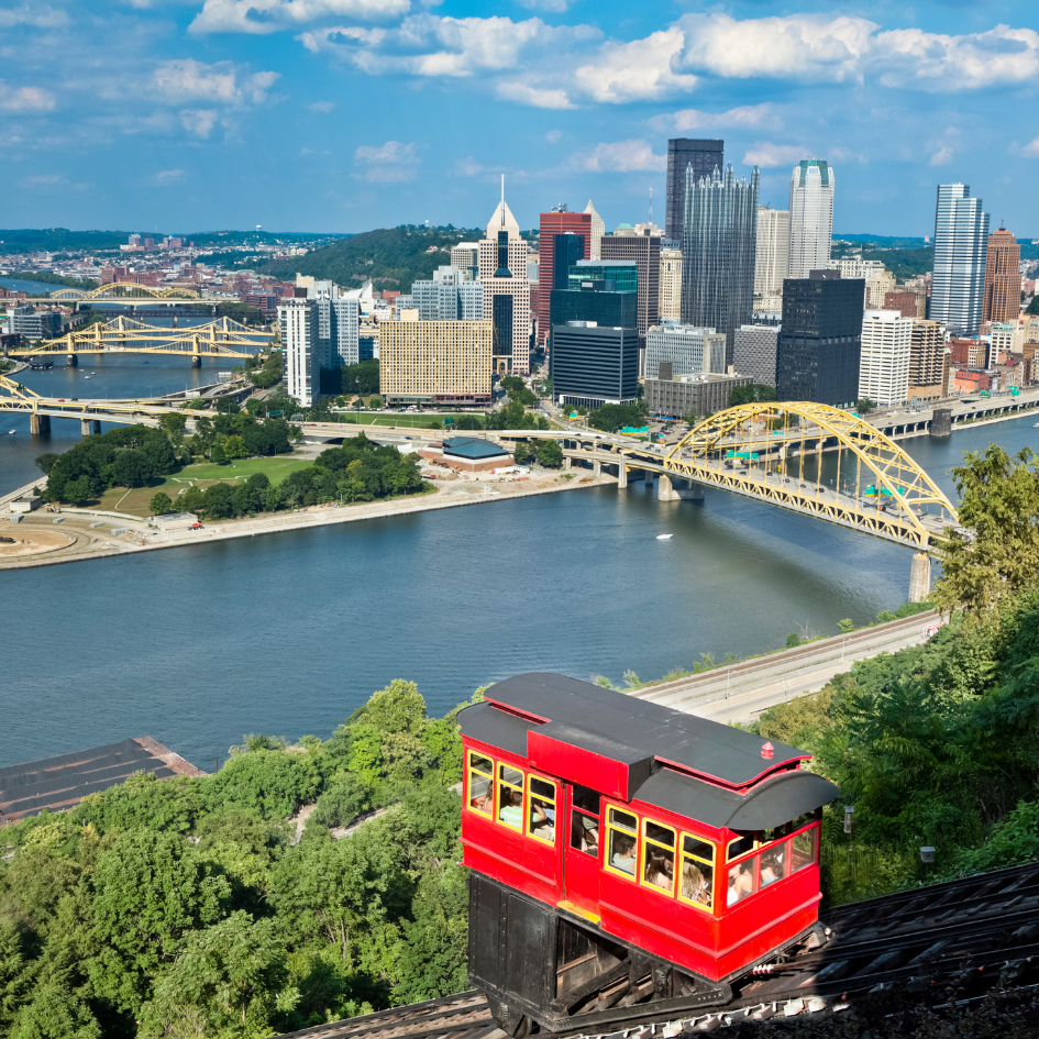 A picturesque view of downtown Pittsburgh with the Duquesne Incline in the foreground, featuring a bright red cable car descending the hillside. The rivers, bridges, and skyline create a stunning backdrop, showcasing the city's unique geography and architecture on a sunny day.