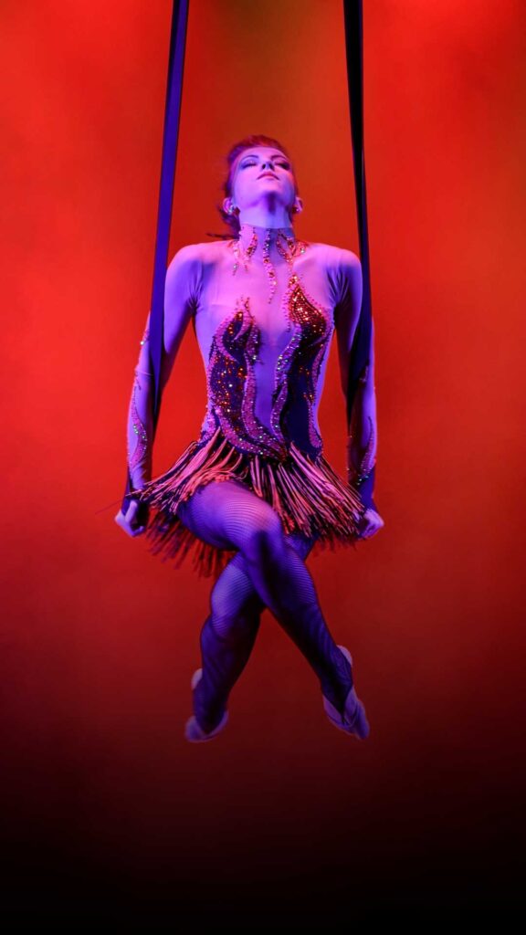 Aerial performer in a vibrant costume suspended mid-air with red and purple lighting in the background