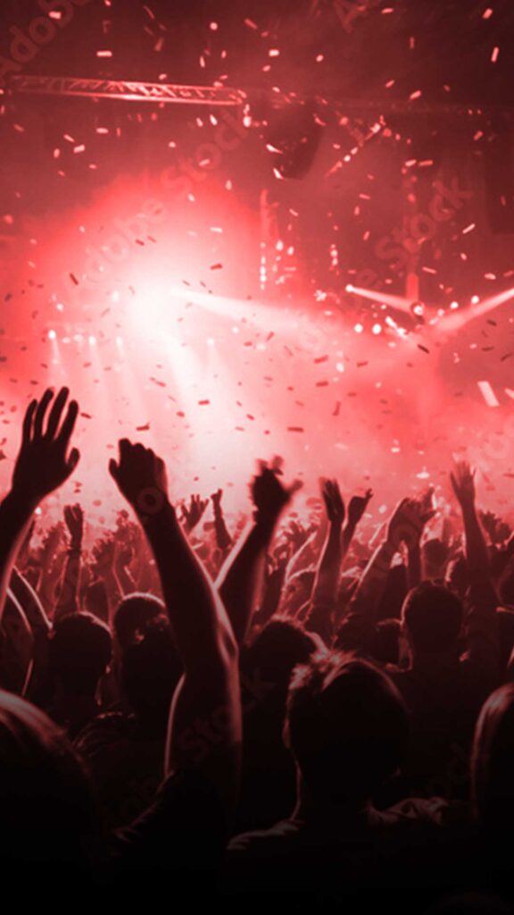 A large crowd with hands raised in the air at a concert, surrounded by confetti and red lighting effects