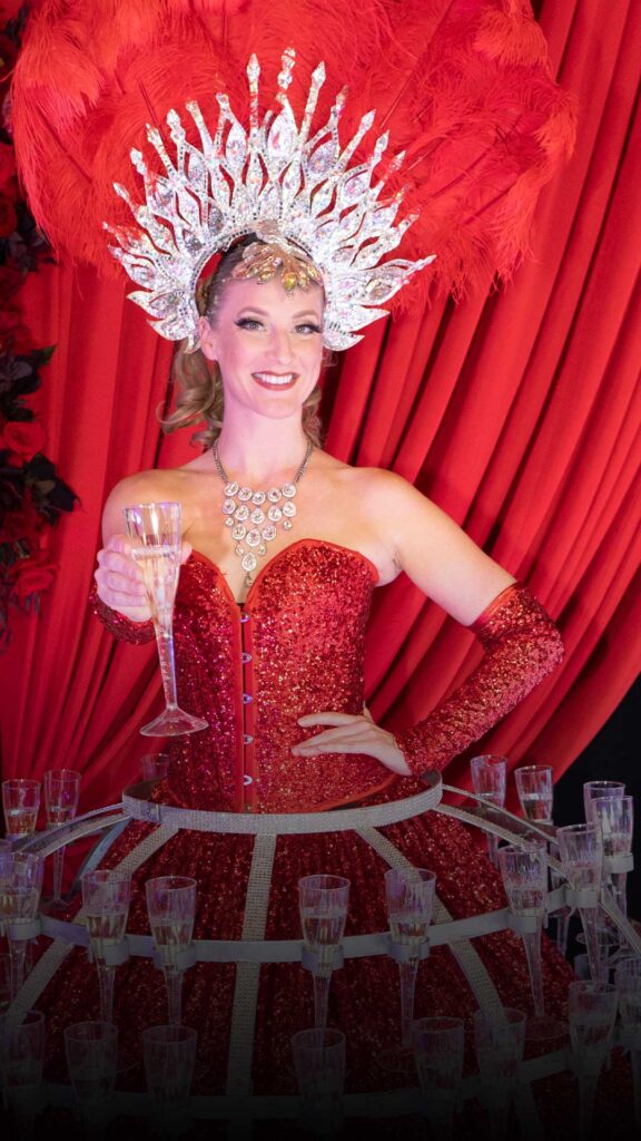 A smiling champagne showgirl dressed in a red sequined outfit with a feathered headdress, holding a glass of champagne