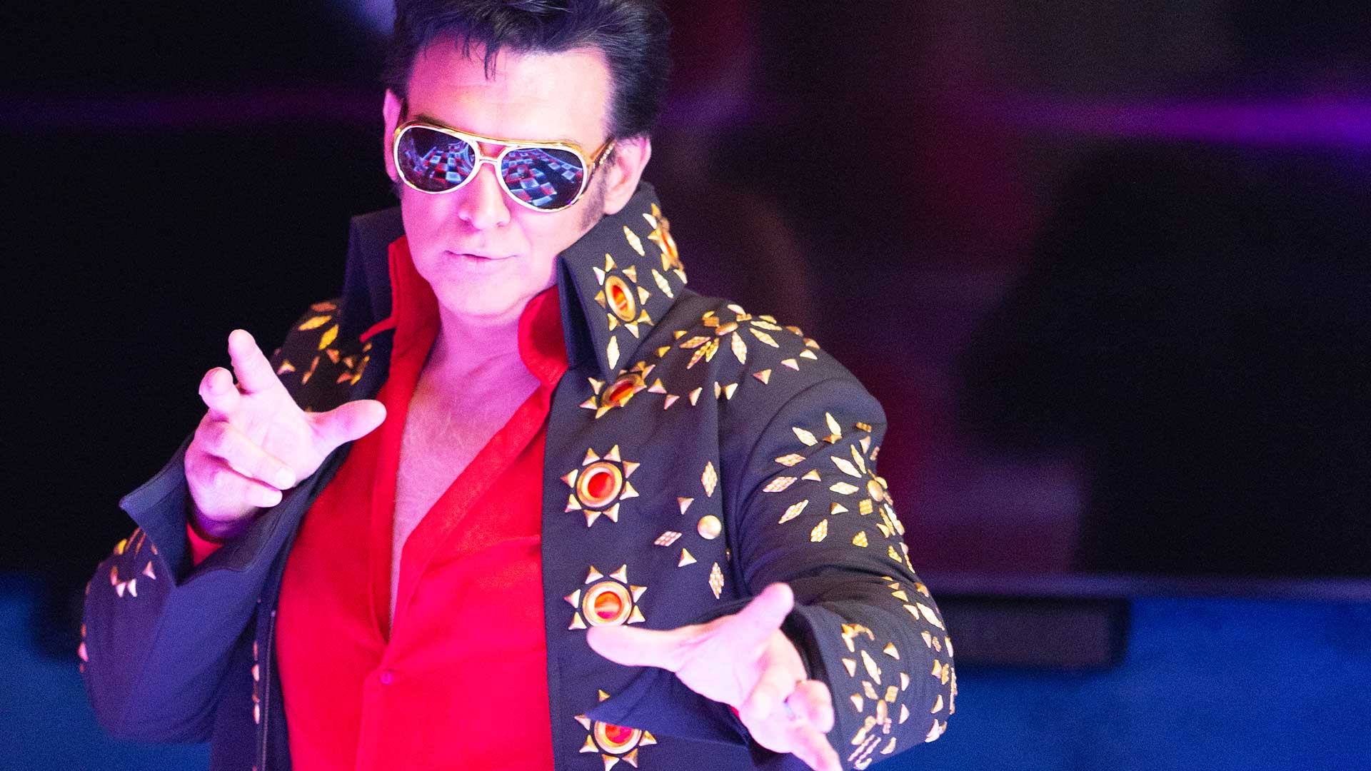 An Elvis impersonator dressed in a red shirt and black studded jacket, performing at a Destination Fabulous event