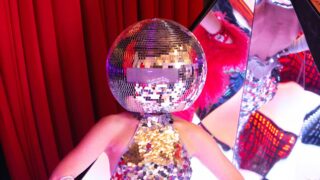 A performer wearing a disco ball helmet and sequin outfit at a Destination Fabulous event, standing in front of a red curtain
