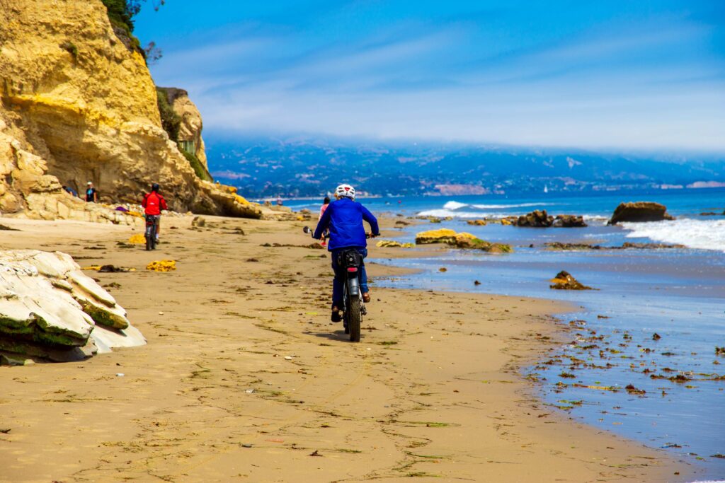a man wearing a red jacket riding an electric bike on the brown sandy beach with people walking along the beach, blue ocean water and lush green trees at Leadbetter Beach in Santa Barbara California