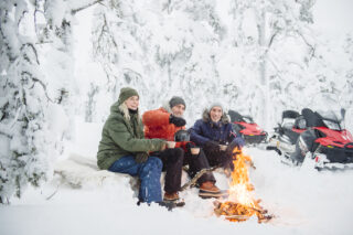 Three people sitting in a snowy setting in front of a campfire.