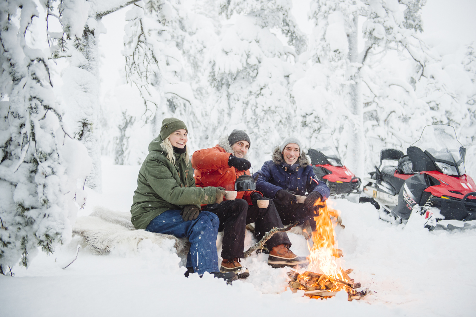 Three people sitting in a snowy setting in front of a campfire.