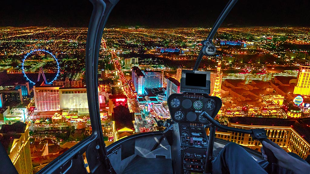 A helicopter cockpit view over the brightly lit Las Vegas Strip at night, showcasing iconic landmarks like the High Roller Ferris wheel.